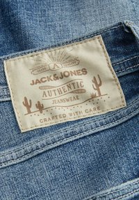 Label from Jack & Jones jeans, tan fabric with printed text and cactus design, attached to blue denim, showing stitching and texture details.