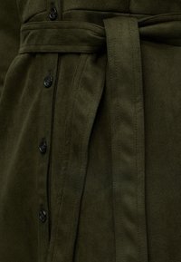Σκούρο πράσινο μάλλινο παλτό με ορατά μαύρα κουμπιά και υφασμάτινη ζώνη δεμένη στη μέση.