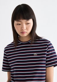 T-shirt rayé marine avec des lignes rouges et bleu clair, encolure ronde et manches courtes, comportant un petit logo de crocodile vert sur la poitrine.
