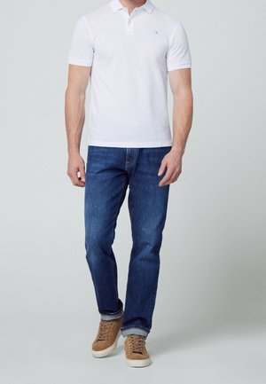 Weißes Polo-Shirt, kurze Ärmel, klassischer Kragen; kombiniert mit dunkelblauen Jeans und hellbraunen Wildledersneakers mit cremefarbenen Sohlen. Schlichtes Design, keine Muster.