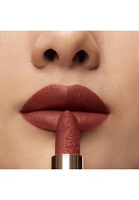 Mat læbestift i en varm, jordagtig brun nuance holdes mellem fyldige læber. Stiften har indviklede indgraveringer og en gylden metalbase.