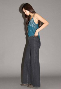 Tyrkysová tielko s vyšívanými detailmi v kombinácii so širokými tmavošedými džínami. Hladká textúra so priliehavým pásom a vzorovanou kvetinovou výšivkou.