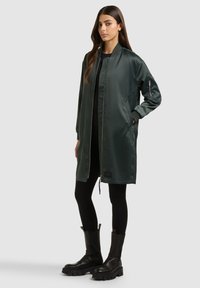 Pikkade pruunide juustega naine kannab tumerohelist pika bomber-jakki, musti retuuse ja musti massiivseid saapaid, seistes ühevärvilise tausta ees.