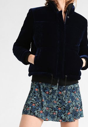 Kobieta nosząca czarną aksamitną pikowaną kurtkę z przednimi kieszeniami oraz krótką kwiatową spódnicę w niebieskie i czerwone kwiaty.