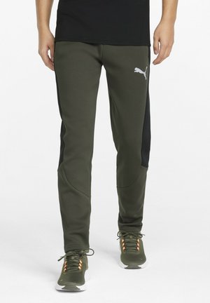 Hombre con pantalones de chándal Puma verde oliva con paneles laterales negros y zapatillas verde oliva a juego con ojales naranjas.