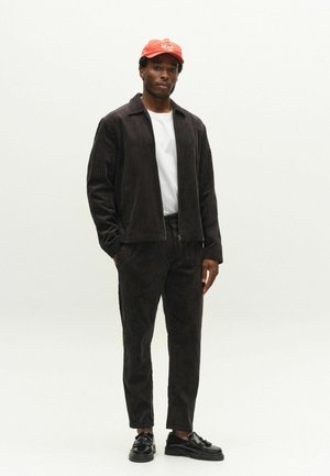 Mann, der eine schwarze Cordjacke und Hose, ein weißes T-Shirt, schwarze Schuhe und eine rote Baseballkappe trägt, steht vor einem schlichten hellen Hintergrund.