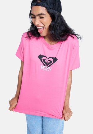 Rosa Baumwoll-T-Shirt mit schwarzem Herz und Roxy-Logo in der Mitte. Locker geschnitten, kurzärmelig, kombiniert mit hellblauem Denim.