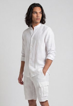 Giovane uomo con capelli scuri di media lunghezza che indossa una camicia bianca con bottoni e pantaloncini cargo bianchi, in piedi con una mano in tasca.