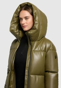 Olijfgroene gewatteerde jas met een grote afneembare capuchon, glanzende textuur, rits aan de voorkant en een zwart geribbeld shirt eronder. Zwarte knoopdetails op de mouwen.