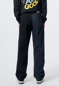 Pantalones de mezclilla negros de pierna ancha con una cintura alta, que cuentan con dos bolsillos traseros y una textura sutil. El modelo lleva zapatillas grises.