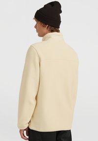 Mann i beige fleece-jakke og svart lue, vendt sidelengs med ryggen mest mot betrakteren på hvit bakgrunn.