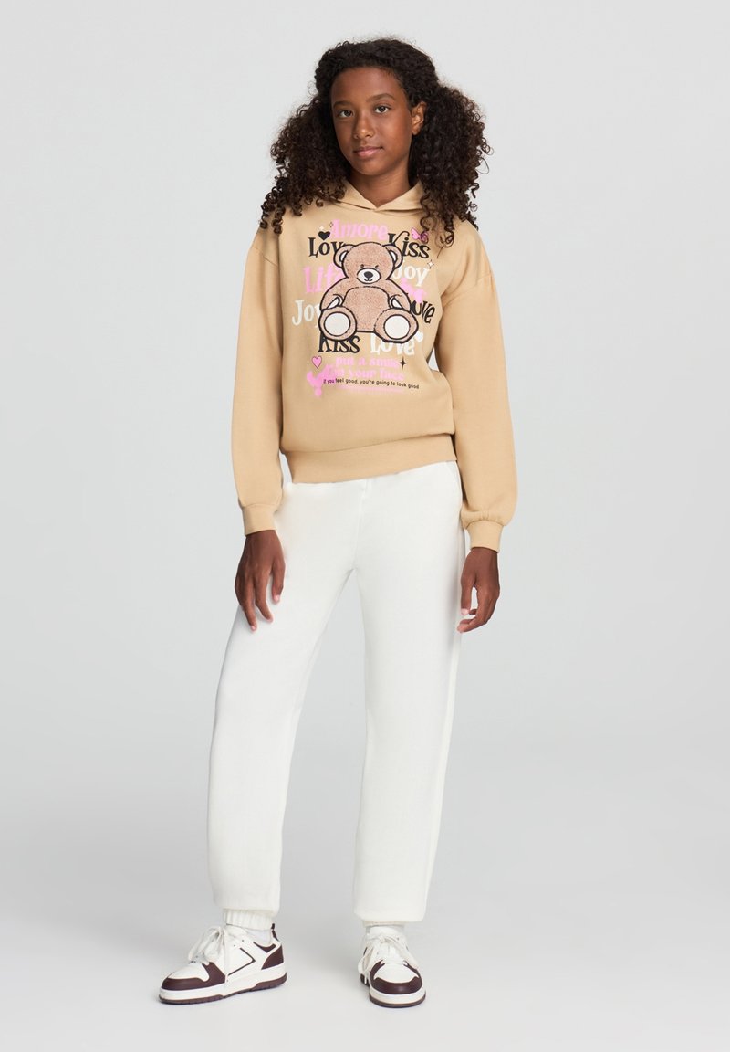 Sweatshirt beige con design di orso grafico, caratterizzato da testo colorato e cuori. Indossato con pantaloni bianchi a sigaretta e sneakers con bordo nero.