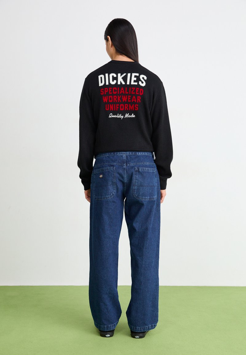 Pull noir avec texte blanc et rouge dans le dos, associé à un jean denim bleu foncé à jambes larges. Design simple et coupe décontractée.