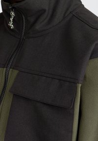 Fleecejack met een hoge kraag, met zwarte en olijfgroene panelen, ritssluiting en een grote borstzak. Gestructureerde stof.