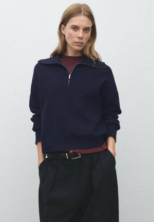 Navy blue quarter-zip sweater in a soft fabric, featuring a collared neckline and relaxed fit, worn over a maroon top and paired with black trousers.