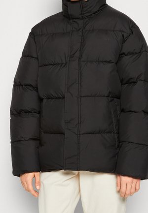 Person trägt eine schwarze gesteppte Steppjacke mit Seitentaschen und cremefarbene Hose, steht vor einem einfarbigen Hintergrund.