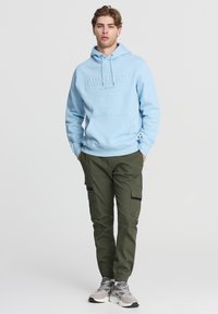 Felpa blu chiaro con logo in rilievo, abbinata a pantaloni cargo verdi oliva con tasche laterali e scarpe da ginnastica grigie.