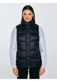 Naine kannab musta puffervesti helehallil dressipluusil, seisab ühevärvilise helehalli tausta ees.