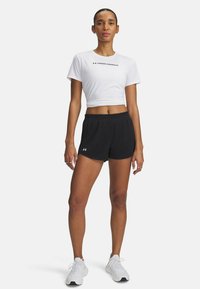 Camiseta atlética blanca corta con logo, combinada con pantalones cortos de malla negros y zapatillas deportivas blancas. El modelo se encuentra en una pose neutral.