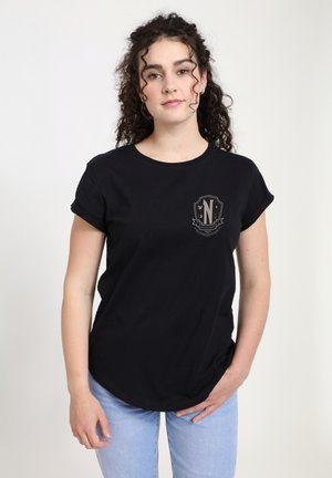 Giovane donna con capelli ricci che indossa una maglietta nera con maniche arrotolate e jeans azzurri chiari, in piedi davanti a uno sfondo bianco uniforme.