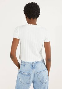 Weißes, geripptes Oberteil mit kurzen Ärmeln und Rüschenausschnitt, kombiniert mit hellblauen, hoch taillierten Jeans mit sichtbaren Gesäßtaschen.