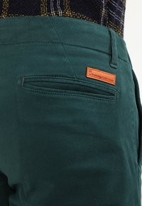 Gröna bomullsbukser med slät front, sidofickor och en brun läderlapp med varumärket "Knowledge Cotton Apparel".