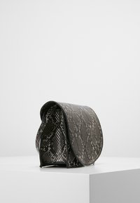 Pequeño bolso negro y gris con textura de piel de serpiente, cierre de solapa y bandolera, exhibido sobre un pedestal blanco contra un fondo liso.