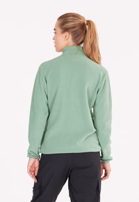 Grüner Fleece-Pullover mit hohem Kragen und langen Ärmeln. Lockerer Schnitt, weiches Material und sorgfältige Nähte, getragen mit schwarzen Hosen.