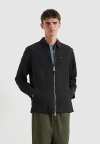 Junger Mann, der eine schwarze Zip-Jacke mit kleinem gelben Logo auf der Brusttasche und olivgrünen Hosen trägt, steht vor einem hellen Hintergrund.