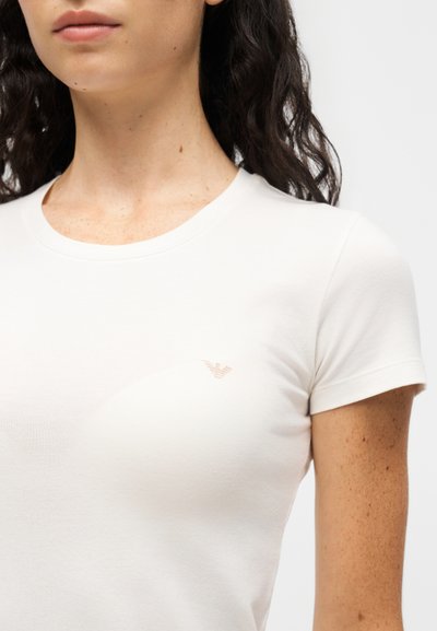 Γυναίκα που φοράει ένα απλό λευκό T-shirt με κοντά μανίκια και μικρό μπεζ λογότυπο στο αριστερό στήθος, εμφανίζεται από τους ώμους μέχρι το πάνω μέρος του στήθους.