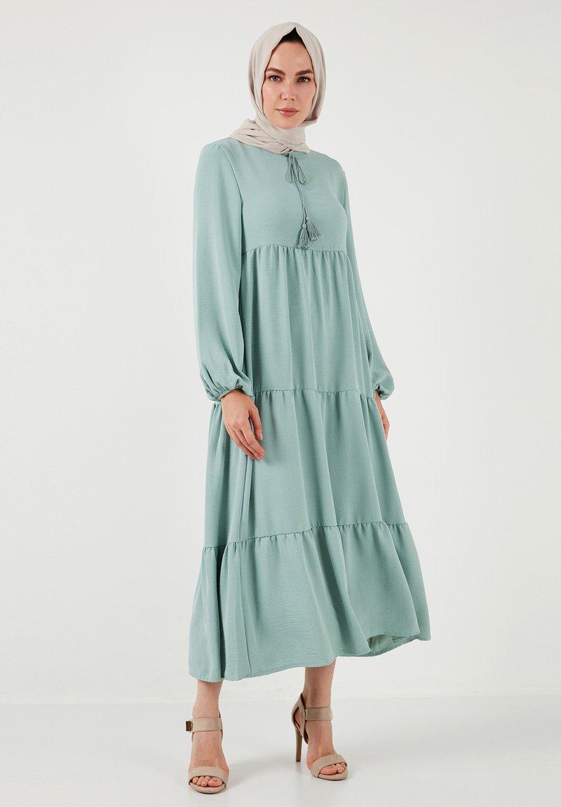 Light green, long-sleeve maxi dress with tiered design, tassel detail at the neckline, and a loose, flowing silhouette. Worn with beige heels.