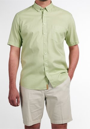 Mann trägt ein hellgrünes kurzärmeliges Hemd mit Tasche und beige Shorts, steht mit einer Hand in der Tasche.