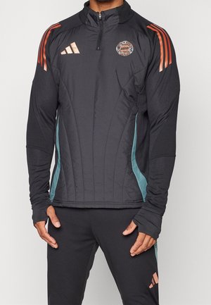 Mann trägt schwarze Adidas-Sportjacke mit orangefarbenen Streifen auf den Schultern und Bayern München-Logo auf der Brust, kombiniert mit schwarzen Hosen.