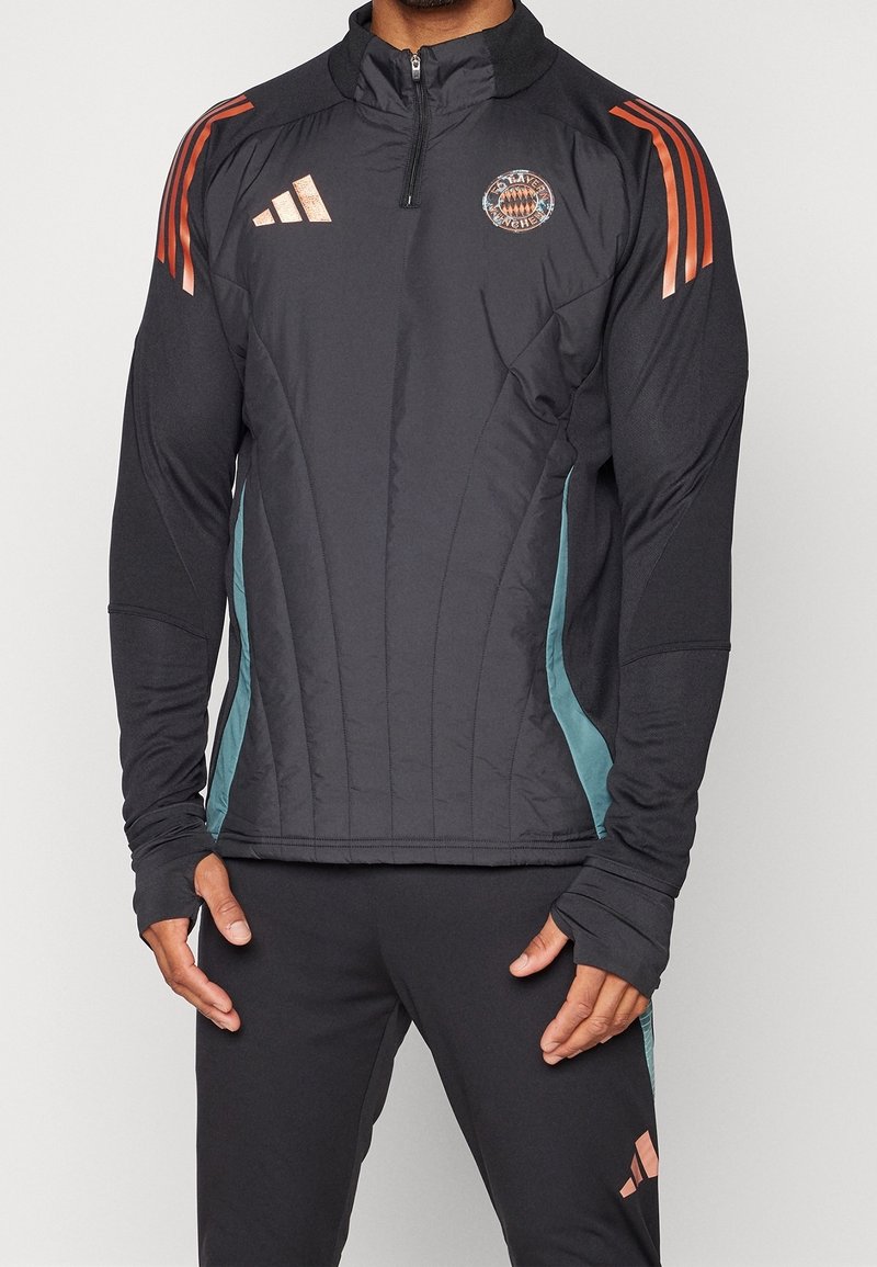 Homme portant une veste de sport Adidas noire avec des bandes orange sur les épaules et le logo du Bayern Munich sur la poitrine, associée à un pantalon noir.