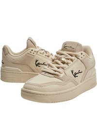 Beige spordijalatsid tekstuurses pinnas, millel on väljapaistav logo, paeltega disain, pehmendusega pahkluudeosa ja dekoratiivsed metallaktsendid.