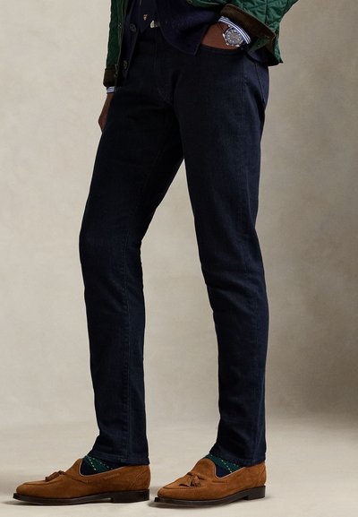 Mørkeblå jeans med smal passform, brune semskede loafers med dusk, og grønne sokker med gule prikkemønster. Teksturert, quiltet jakke synlig.