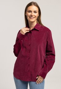 Czerwona koszula z długim rękawem z miękkiego materiału kordurowego, z wyłogami, długimi rękawami i zaokrąglonym dołem; zestawiona z niebieskimi dżinsami.