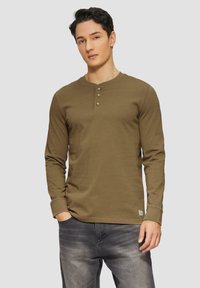 Camicia henley a maniche lunghe color khaki con placca a tre bottoni e polsini a costine, abbinata a jeans grigi scuri. Tessuto dalla finitura liscia e strutturata.