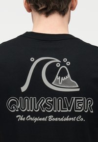 Πίσω μέρος μαύρου πουκάμισου με μεγάλο γκρι στυλιζαρισμένο κύμα και λογότυπο με βουνό λάβας και κείμενο "QUIKSILVER The Original Boardshort Co.".