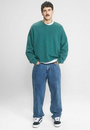 Mężczyzna z wąsami, ubrany w turkusową bluzę, szerokie niebieskie dżinsy i czarno-białe sneakersy, stoi z rękami w kieszeniach na białym tle.