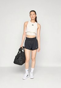 Egy nő fehér, rövid szabású tank toppban és fekete rövidnadrágban áll, egy fekete tornazsákot tart a kezében, fehér Adidas sportcipőt és zoknit visel, egy sima háttér előtt.