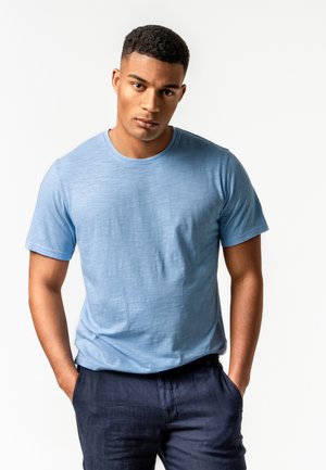 Junger Mann mit kurzen schwarzen Haaren, der ein hellblaues T-Shirt und dunkle Hosen trägt, steht mit den Händen in den Taschen vor einem weißen Hintergrund.