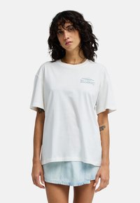 Mujer con cabello rizado y oscuro, vestida con una camiseta blanca de Billabong y una falda vaquera azul claro, sobre un fondo liso.