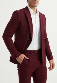 Costume bourgogne à coupe ajustée, veste à un bouton, chemise blanche et accents en métal doré aux manches. Texture lisse et élégante.