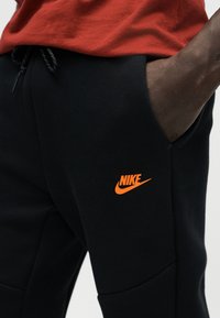 Άτομο που φοράει μαύρο παντελόνι φόρμας Nike με πορτοκαλί λογότυπο και κόκκινο πουκάμισο, το χέρι στην τσέπη, κοντινό στο ύψος της μέσης και του άνω μέρους του ποδιού.