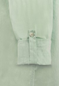 Puño de camisa de lino verde claro con un solo botón de cierre, textura ligeramente fruncida y un acabado suave a lo largo de la manga.