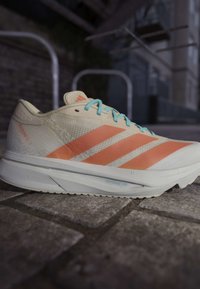 White and orange Adidas running shoe with blue laces placed on stone pavement in an outdoor urban setting.