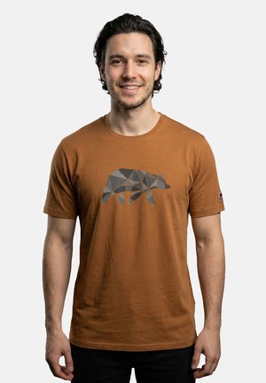 Mann mit dunklen Haaren, der ein braunes T-Shirt mit einem geometrischen Bärendesign trägt, steht vor einem einfarbigen hellgrauen Hintergrund.