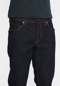 Jean en denim bleu foncé avec des coutures jaunes, design à cinq poches et fermeture à bouton métallique à la taille. Forme de jambe légèrement fuselée.