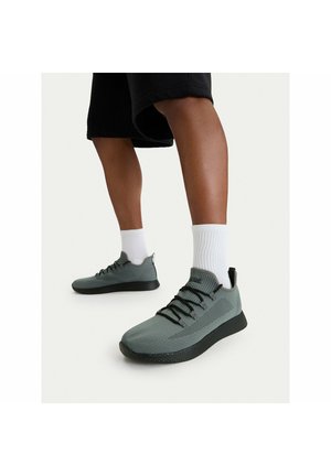 Osoba stojící na bílém povrchu, obutá v šedých pletených teniskách s černými podrážkami, bílé ponožky a černé šortky.